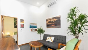 Una sala de estar moderna con un sofá gris, una mesa de centro de madera y un sillón verde. En la pared, hay un cuadro de un atardecer, una planta en maceta y una pequeña mesa con una lámpara.