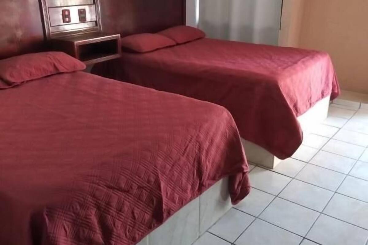 Ein Hotelzimmer mit zwei Betten, einem Holzkopfteil, einem Nachttisch und einem Fenster mit Vorhängen.