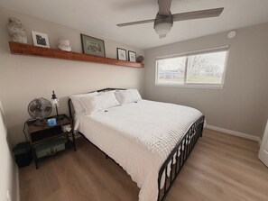 Un dormitorio con cama, ventilador de techo, ventana y estante con fotografías y una lámpara.