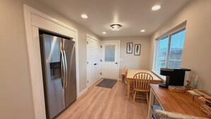 A modern kitchen with a stainless steel refrigerator, wooden dining table, and chairs, a small island with a coffee machine, and a window with a view.