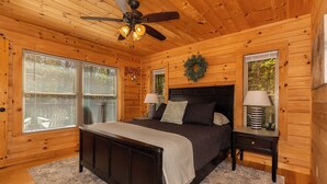 Une chambre dans une cabane en bois, avec un lit, deux tables de chevet, un ventilateur de plafond et une fenêtre avec des stores.