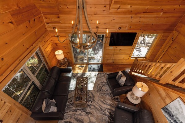 L’intérieur d’un chalet en bois chaleureux et accueillant, avec un canapé en cuir noir, un lustre et une grande fenêtre donnant sur le paysage extérieur.