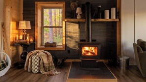 L’intérieur chaleureux d’un chalet avec un poêle à bois, une fenêtre donnant sur la forêt, une lampe et un plaid à carreaux sur le sol.