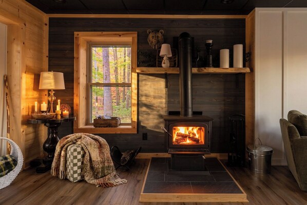 Interior acogedor de una cabaña con estufa de leña, ventana con vista al bosque, lámpara y manta de cuadros en el suelo.