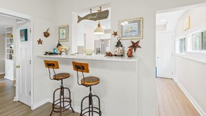 Een keuken met een wit aanrecht, twee houten barstoelen, een hanglamp en decoratieve elementen zoals een bord met een vis en zeesterren.