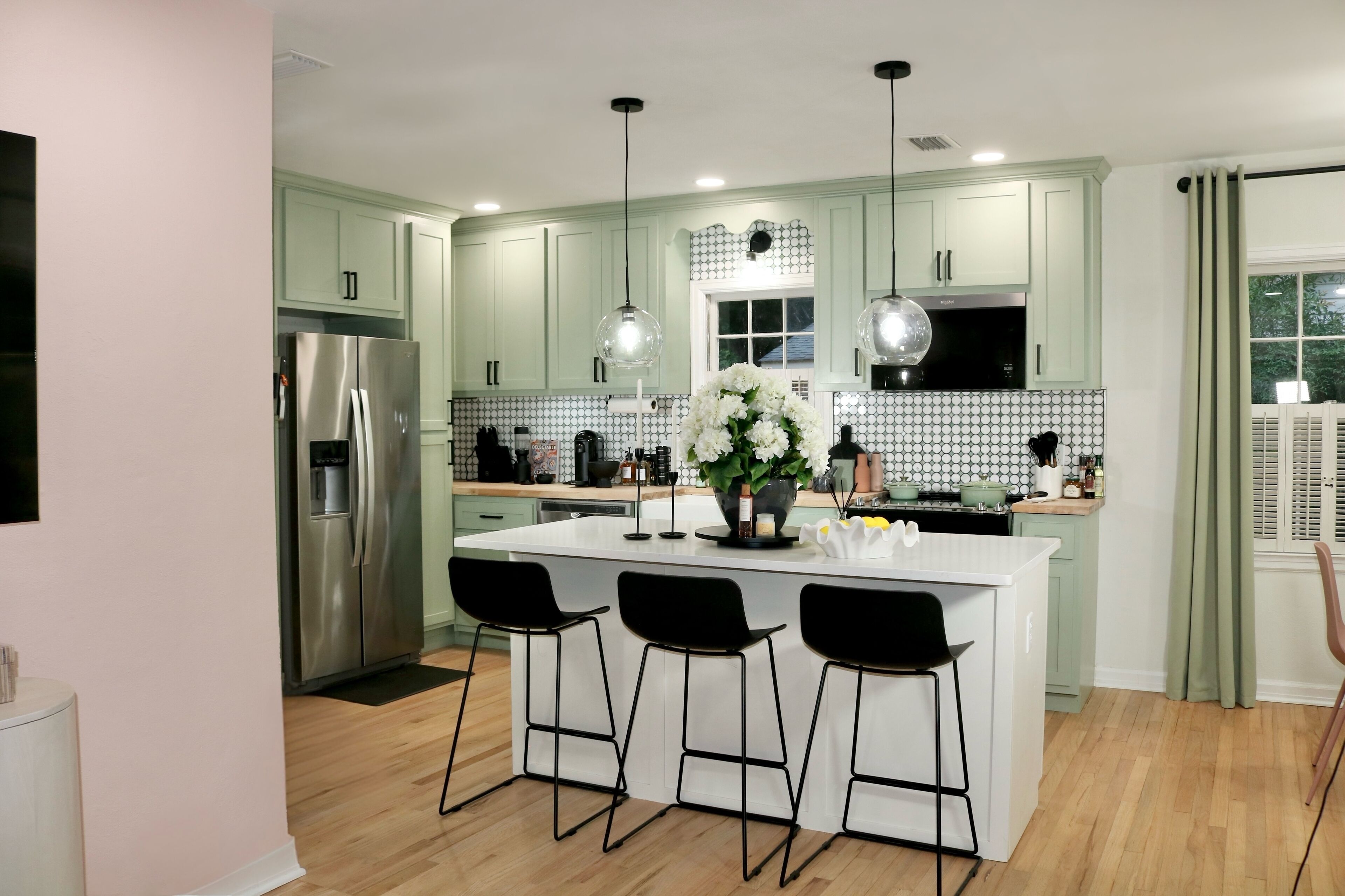 Een moderne keuken met groene kasten, een roestvrijstalen koelkast, een centraal kookeiland met barkrukken, hanglampen en een decoratieve bloemstelling.