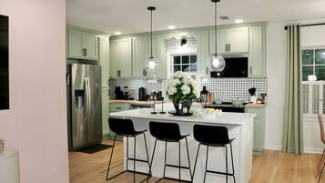 Una cocina moderna con gabinetes verdes, un refrigerador de acero inoxidable, una isla central con taburetes de bar, luces colgantes y un arreglo floral decorativo.