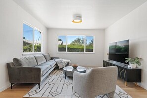 Una sala moderna con un sofá gris, una mesa redonda para café, un televisor sobre un mueble negro y un ventanal con vistas a zonas verdes.