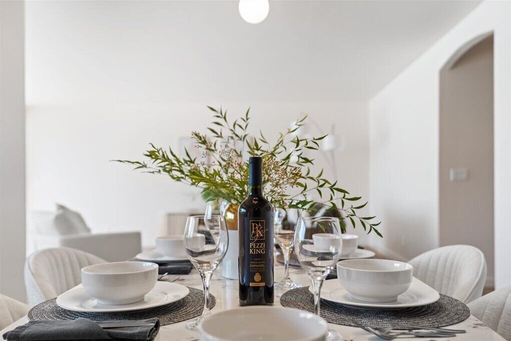Mesa de comedor preparada con platos, fuentes y copas de vino, centrada en una botella de vino.