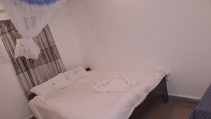 Ein Einzelbett mit weißer Bettwäsche und einem herzförmigen Kissen, einem kleinen Teppich und einer karierten Tagesdecke auf einem hölzernen Bettgestell in einem Zimmer mit schlichten Wänden und einem Vorhang mit Muster.