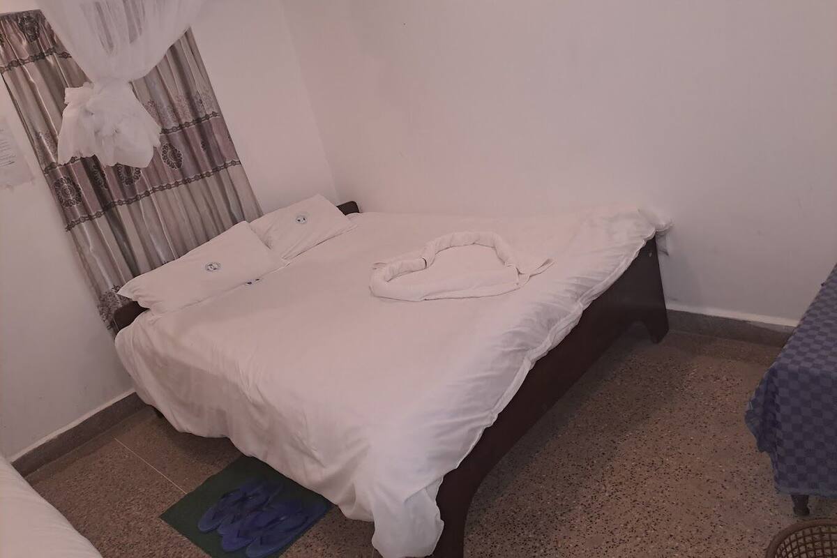 Una cama individual con ropa de cama blanca, un cojín con forma de corazón, una alfombra pequeña y una colcha a cuadros sobre un cabecero de madera en una habitación con paredes lisas y una cortina estampada.
