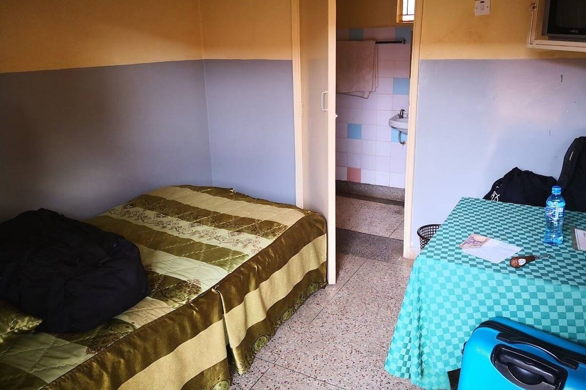 Una habitación pequeña y funcional con una cama individual, una mesa con mantel a cuadros y un televisor montado en la pared.