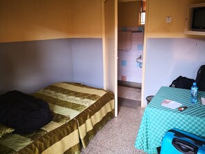 Une petite pièce, de taille modeste, comprenant un lit simple, une table recouverte d’une nappe à carreaux et un téléviseur fixé au mur.
