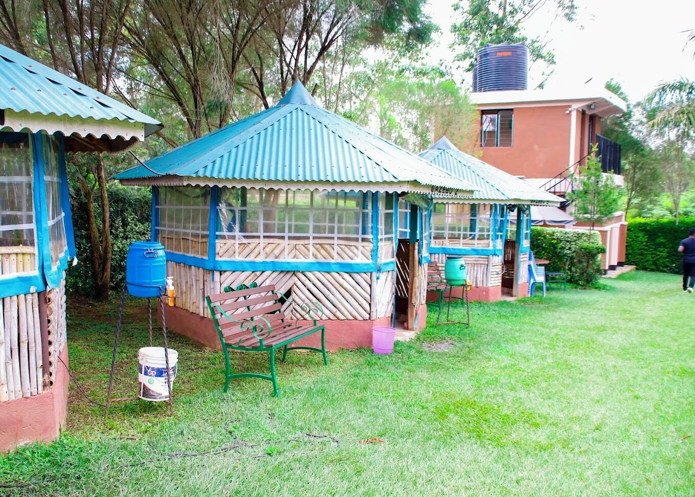 A rustic outdoor seating area with wooden benches and a covered structure, surrounded by greenery and a building with a water tank on the roof.