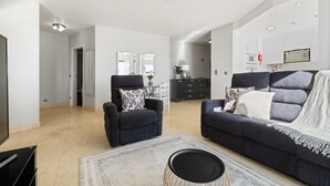 Ein modernes Wohnzimmer mit einer dunklen Couch, einem Sessel und einem Couchtisch. Im Hintergrund ist eine Küchenzeile mit weißen Schränken und einer Mikrowelle zu sehen.