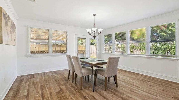 Une salle à manger avec un plancher en bois, un lustre et de grandes fenêtres équipées de stores.