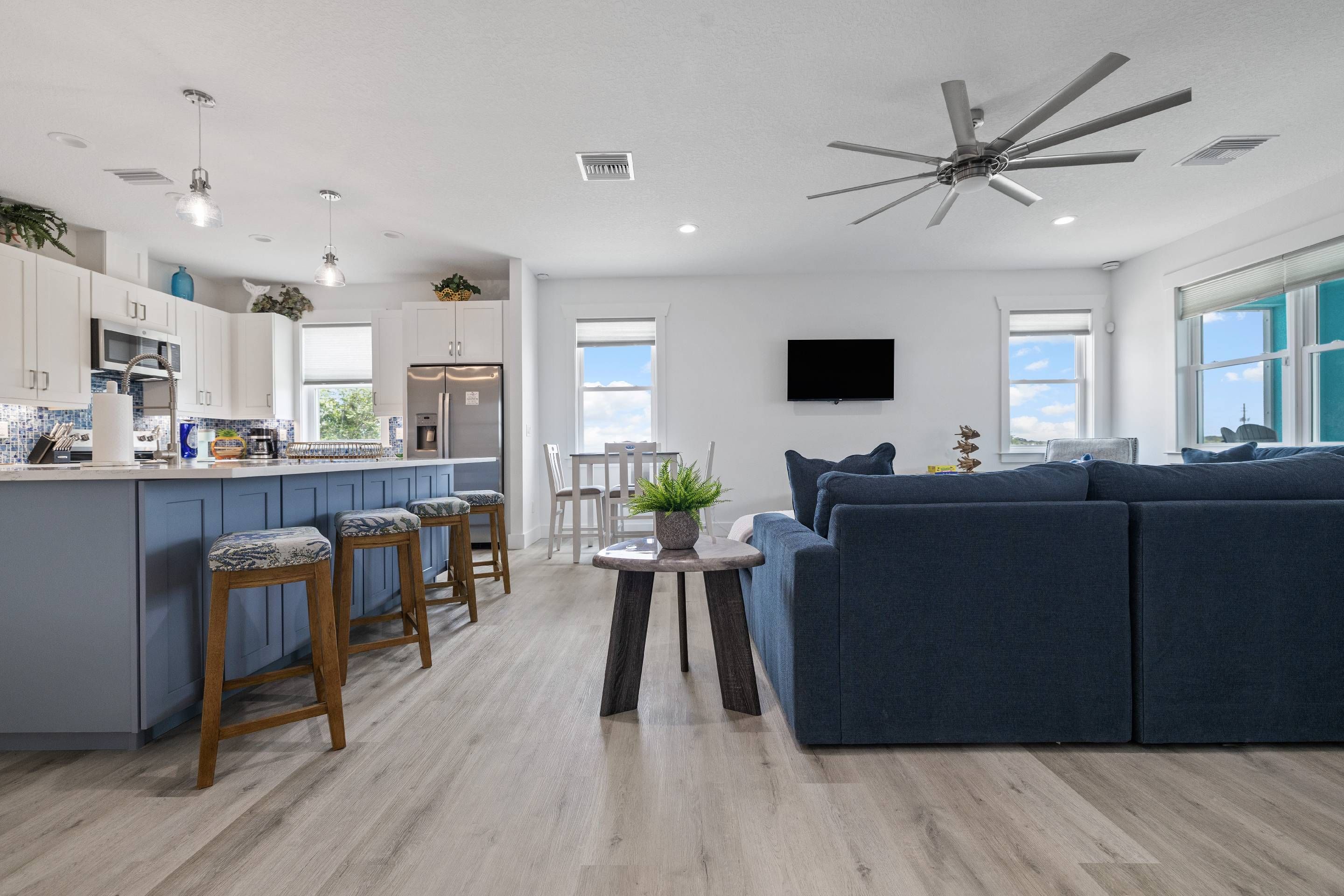 A modern kitchen with a bar area, a dining table with chairs, a blue sofa, and a ceiling fan.