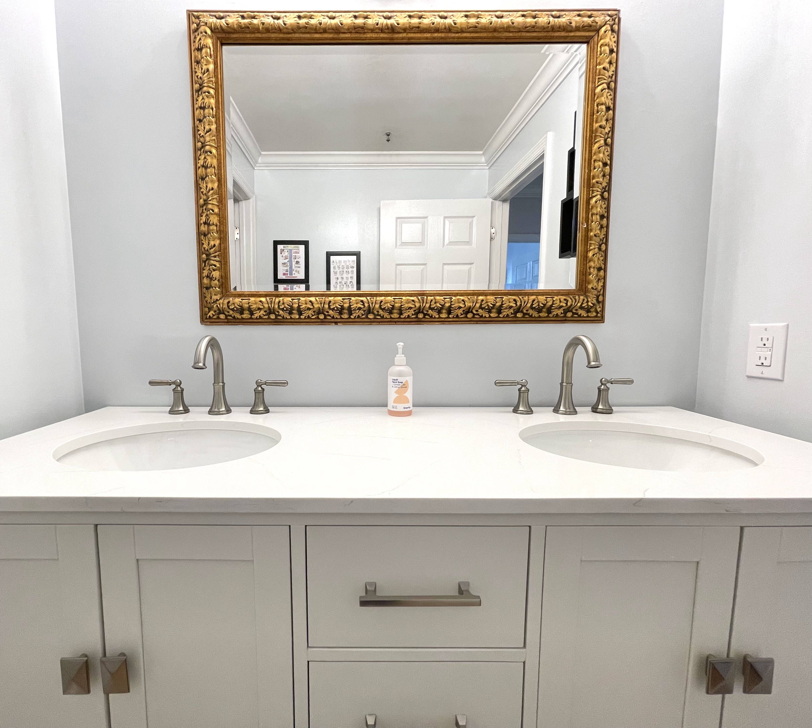 A bathroom with two sinks, a large mirror with an ornate frame, and a bottle of lotion on the countertop.