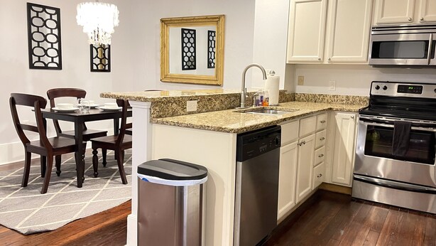 Una cocina moderna con armarios blancos, encimera de granito, electrodomésticos de acero inoxidable y un comedor con mesa y sillas de madera.