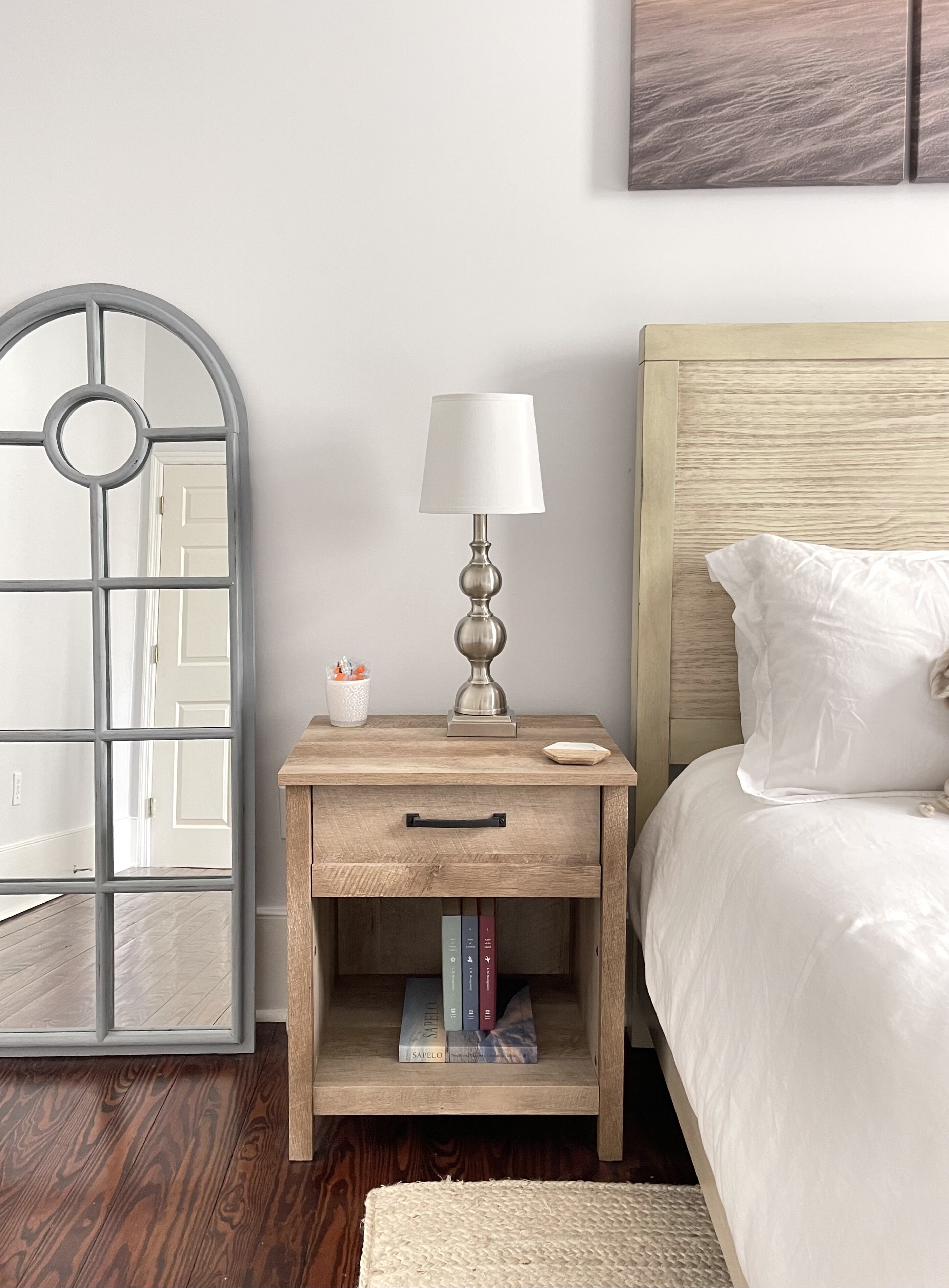 Een slaapkamer met een houten nachtkastje, een lamp, een spiegel en een bed met witte beddengoed.