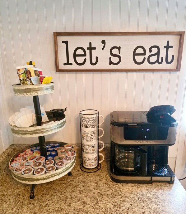 A kitchen counter with a coffee maker, a mug, a tiered serving tray with food items, and a sign that reads 'let's eat'. - Cozy Cottage 1bed 1bath in Slocomb Al (Slocomb)