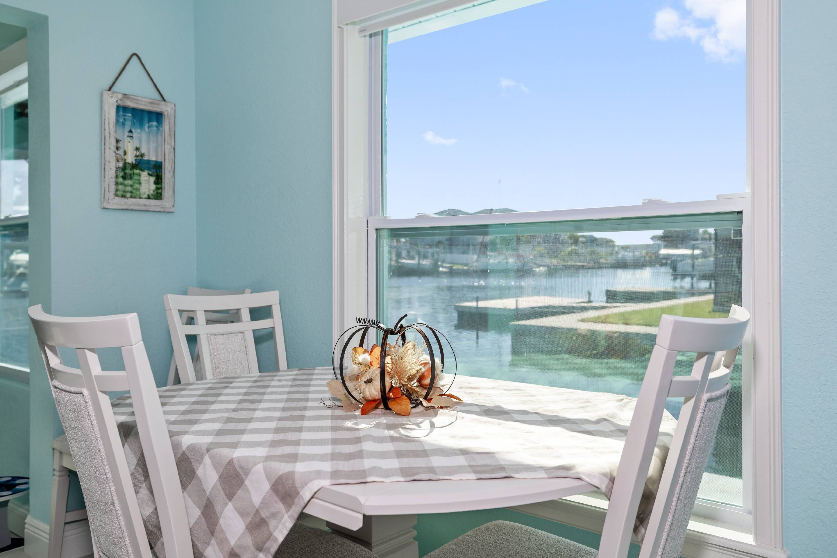 Une table à manger recouverte d’une nappe à carreaux, avec des chaises blanches et un centre de table décoratif. Un tableau encadré est accroché au mur.