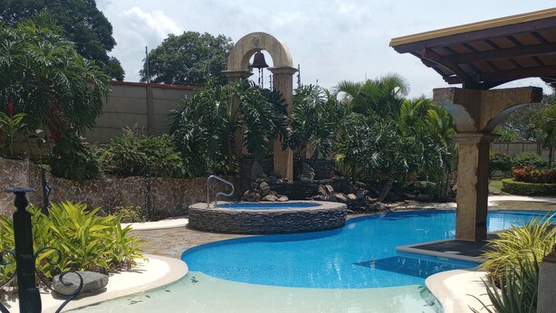 A swimming pool with a stone archway and a bell, surrounded by lush greenery and a stone wall. - La mansión  (San Isidro)