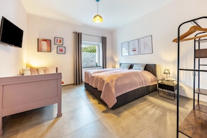 A bedroom with a bed, bedside table, lamp, TV, and a window with curtains. - Suites at the Historic Berghotel Johanna (Eslohe (Sauerland))