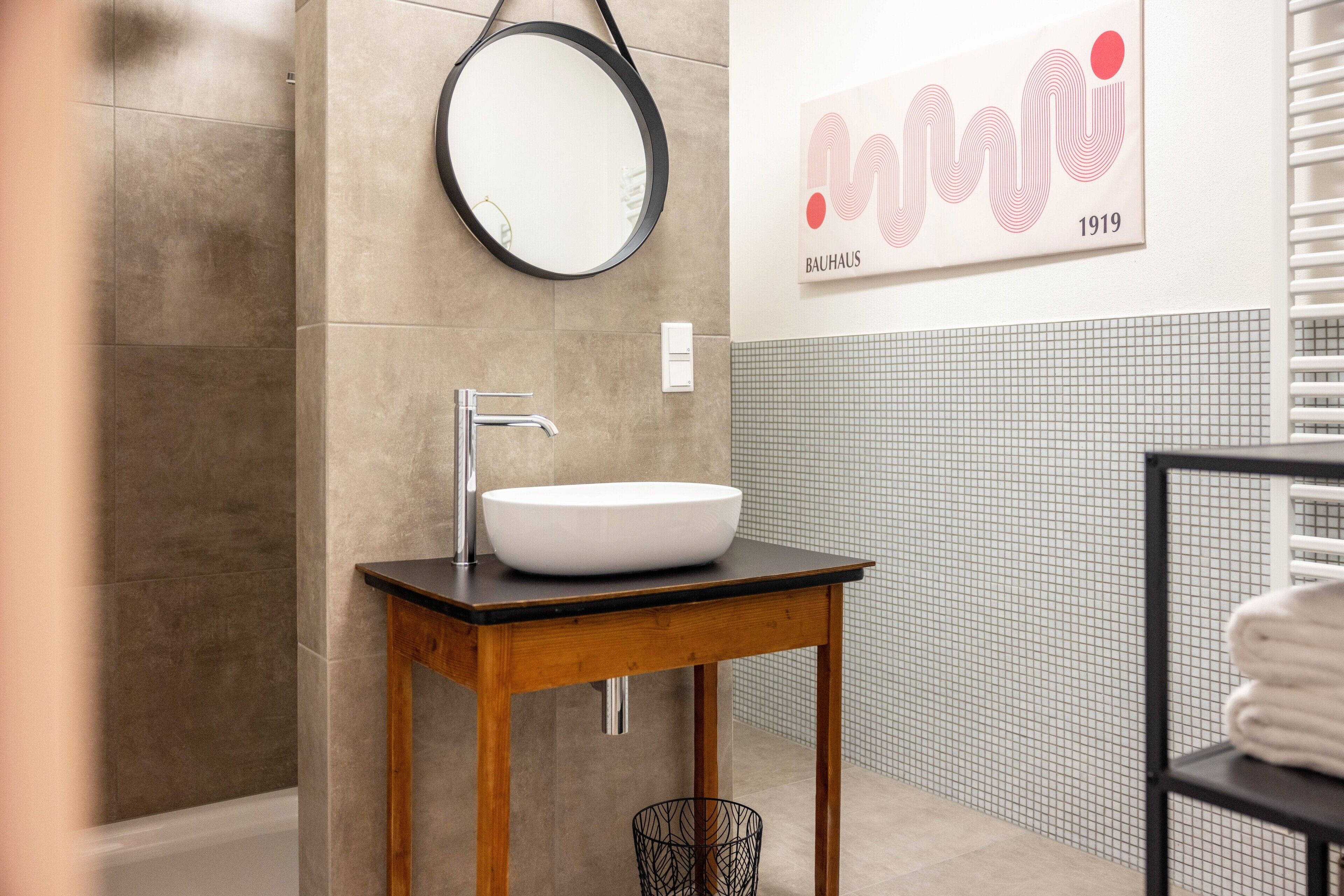 A bathroom with a round mirror, a white sink on a wooden table, a towel rack, and a Bauhaus artwork.