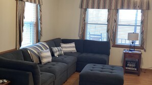 Ein Wohnzimmer mit Ecksofa, Hocker, Deckenventilator und einem Tisch mit einem Schild.