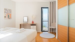 Ein Hotelzimmer mit zwei Betten, einem Schreibtisch, einem Sessel, einem kleinen Rundteppich und Meerblick durch eine Glastür.