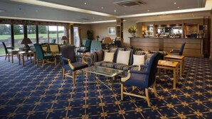 A hotel lobby with a patterned carpet, seating area with sofas and chairs, a bar area, and large windows offering a view of the outdoors. - Dale Hill Hotel (Ticehurst)