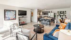 Ein modernes Wohnzimmer mit einer Couch, Sesseln, einem Fernseher und einer Küchenzeile mit Barhocker.