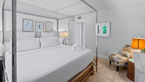 A four-poster bed with white linens, a bedside table with a lamp, and a chair with a patterned cushion.