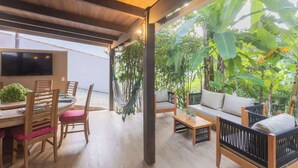 A spacious patio with a wooden dining table, chairs, and a sofa. There is a TV mounted on the wall and a hammock hanging between the chairs.