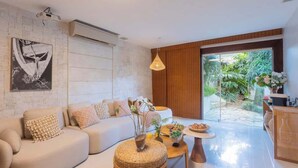 A modern living room with a sofa, a coffee table, and a dining area. There is a large window providing a view of the outdoors.