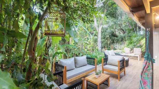 A cozy outdoor seating area with wicker furniture, surrounded by lush greenery and a hanging birdcage.