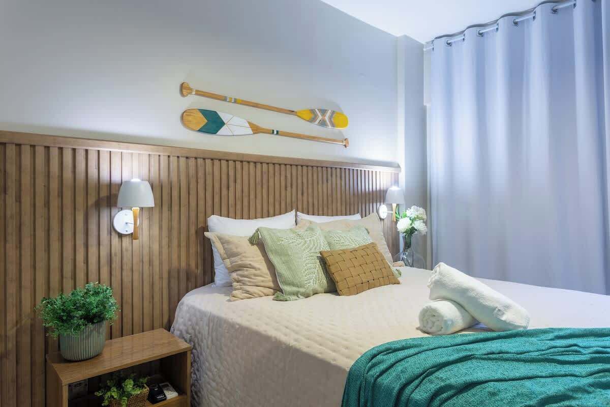Une chambre d’hôtel avec une tête de lit en bois, un lit recouvert d’une couverture verte, une table de chevet ornée d’une plante et une planche de paddle fixée au mur.