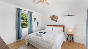 Une chambre à coucher comprenant un lit, un ventilateur de plafond, un appareil de climatisation, une table de chevet avec une lampe et une fenêtre avec des rideaux.