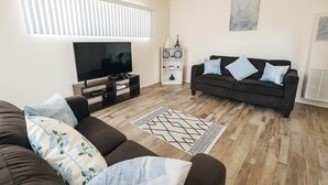 A modern living room with a flat-screen TV, a dark brown sofa with patterned cushions, a wooden floor, and a decorative rug.