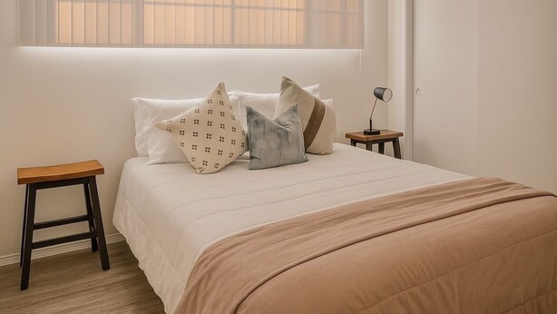 A bedroom with a bed, pillows, a bedside table, a lamp, and a window with blinds.