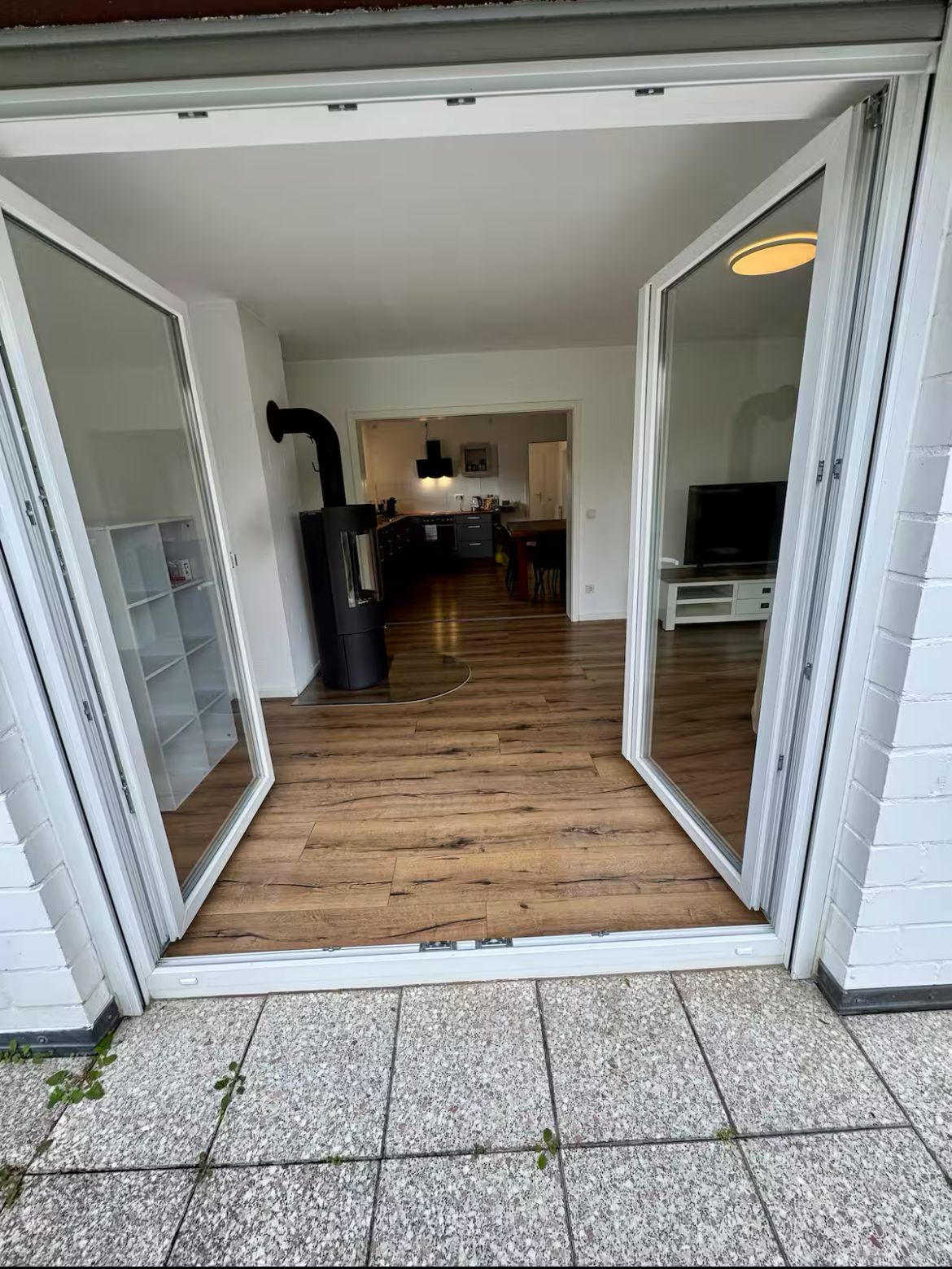 An open sliding glass door leading into a kitchen with a black range hood, wooden flooring, and a dining area with chairs and a table.