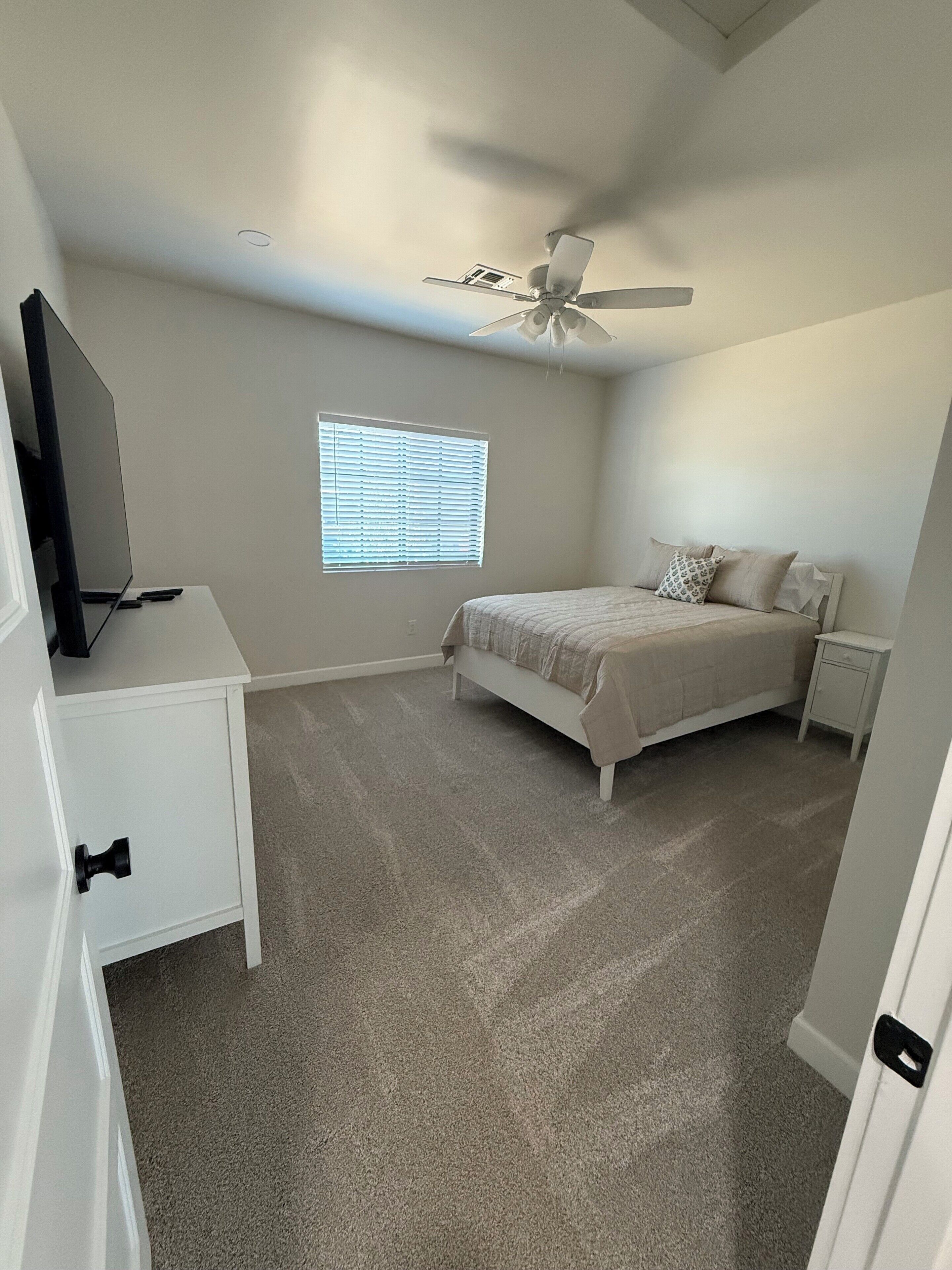 A bedroom with a bed, a TV on a stand, a ceiling fan, a window with blinds, and a carpeted floor.