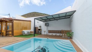 A swimming pool with a logo, a covered patio, a wooden deck, and a mountain in the background. - Vacation Villa 'Montaña' with Wi-Fi and Air Conditioning (Balcon de Telde)