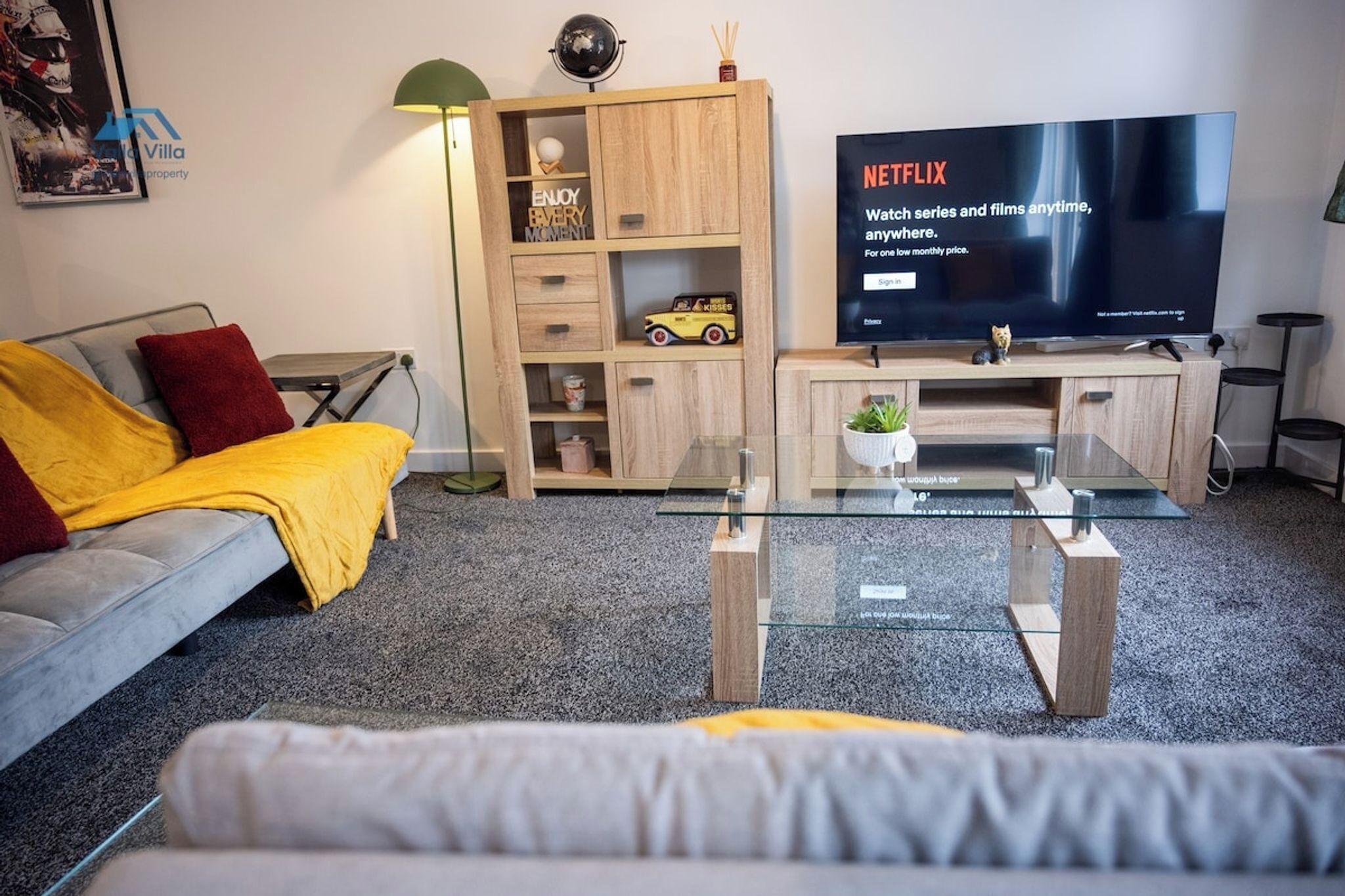 A modern living room with a grey sofa, a yellow throw blanket, a wooden TV stand with a flat-screen TV displaying Netflix, and a glass coffee table.