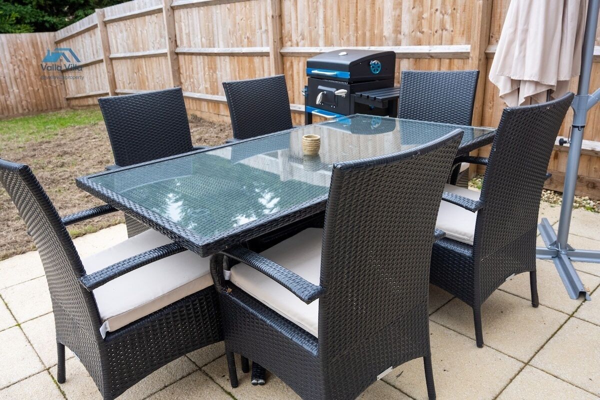 A glass-top table with wicker chairs, a barbecue grill, and a wooden fence in the background.