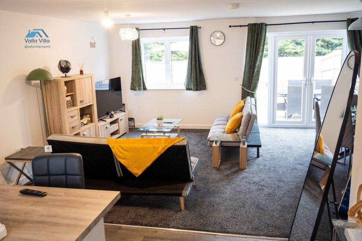 Un salon moderne avec un canapé, une table basse et un téléviseur. On y trouve également une bibliothèque, un bureau et une chaise. La pièce est dotée d’une grande fenêtre avec des rideaux et offre une vue sur la verdure à l’extérieur.