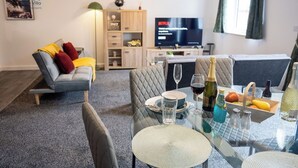 Una sala moderna con un sofá, una mesa de comedor preparada para dos personas, un televisor y una estantería.