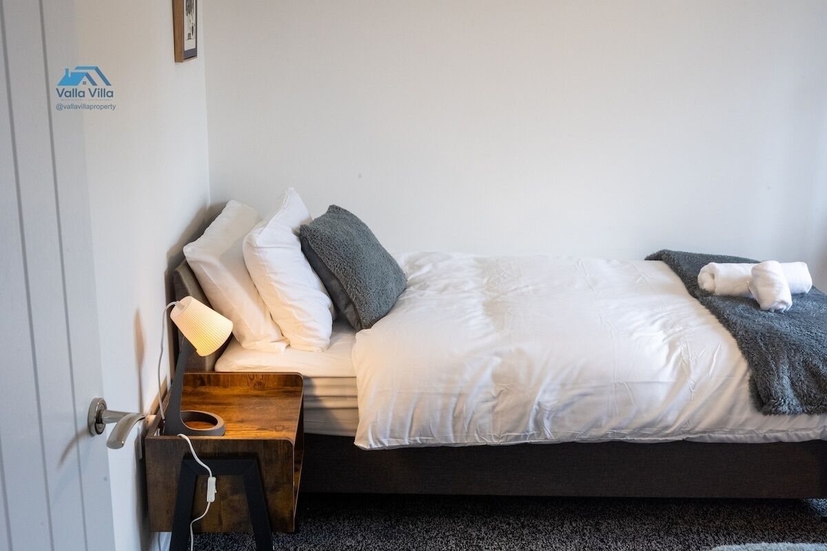 A neatly made bed with white and gray bedding, a bedside lamp, and a wooden nightstand with a charging cable.