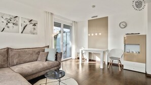 A modern living room with a brown sofa, a round coffee table, and a white rug. There is a white dining table with chairs, a white wall clock, and a window with curtains. - Markgraf Apartments (Bad Bellingen)